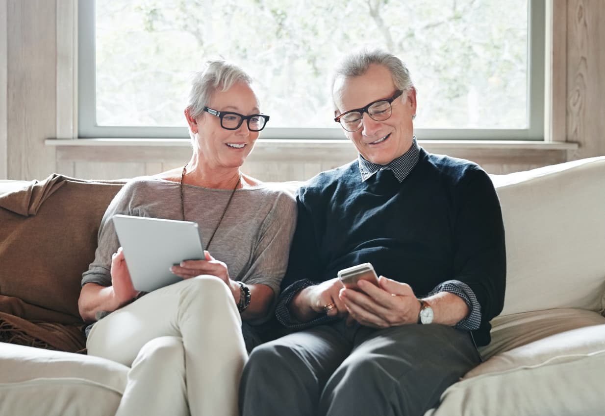 Senior couple sitting on couch and smiling at mobile devices.